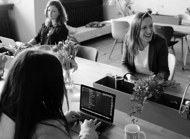 Three women working on a laptop computer
         in an office, one of them laughing with someone offscreen